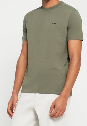 Mann trägt olivgrünes T-Shirt mit kurzen Ärmeln und Rundhalsausschnitt mit „BOSS“-Logo auf der Brust und weißen Hosen, steht vor einfarbigem Hintergrund.