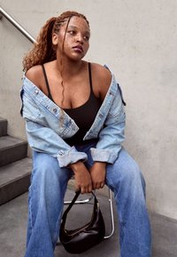 Ženska z pletenicami, oblečena v črn zgornji del oblačila in denim jakno brez ramen, sedi na stolu in drži majhno črno torbico.