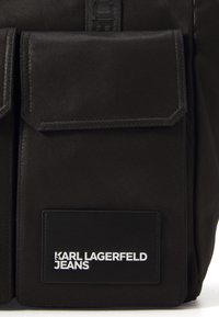 Svart tygryggsäck med två framfickor med lock, en gummilogg med texten "KARL LAGERFELD JEANS" och hållbara stygndetaljer.