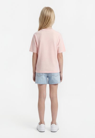 Enfant blond portant un t-shirt rose clair à manches courtes, un short en jean bleu clair et des baskets blanches, de dos sur un fond blanc.
