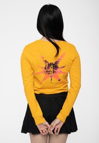 Long-sleeve yellow cotton shirt with a graphic design featuring a black and pink starburst on the back. Paired with a black skirt.