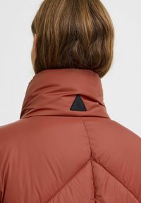 Padded Jacke in Rostfarbe mit hohem Kragen. Verfügt über ein dreieckiges schwarzes Logopatch und diagonale Nähte auf der Rückseite. Glatte Textur.