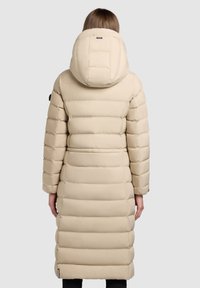 Manteau matelassé beige avec capuche oversize, motif matelassé, longueur longue, manches longues et poches latérales. Texture lisse et construction robuste.