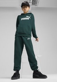 Zielona bluza z kapturem z białym logo "PUMA", w zestawie z dopasowanymi spodniami dresowymi i czarnymi sneakersami. Model nosi czarną czapkę i okulary, stoi obok krzesła.