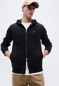 Jeune homme portant une casquette beige, un sweat à capuche noir zippé avec logo, une chemise blanche et un pantalon marron, debout les mains dans les poches du sweat.