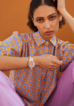 Woman with curly hair wears a patterned orange, white, and blue shirt, pink pants, and a rose gold wristwatch, sitting against an orange background.