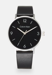 Montre noire avec un boîtier en acier inoxydable rond, des aiguilles et des chiffres argentés. Dispose d'un bracelet en cuir noir à la texture lisse.