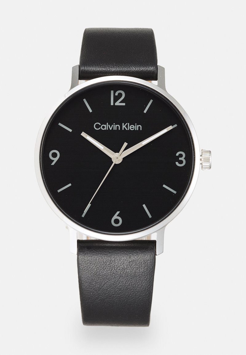 Reloj negro con una caja redonda de acero inoxidable, manecillas y números plateados. Presenta una correa de cuero negro con una textura suave.