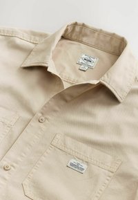 Chemise beige à manches courtes en mélange de coton, dotée d'un col boutonné, d'une poche poitrine et d'une étiquette avec les informations de marque et d'entretien.