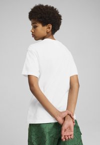 Hvid bomuld t-shirt med korte ærmer og rund hals, båret med grønne mønstrede shorts. Personen vises fra ryggen.