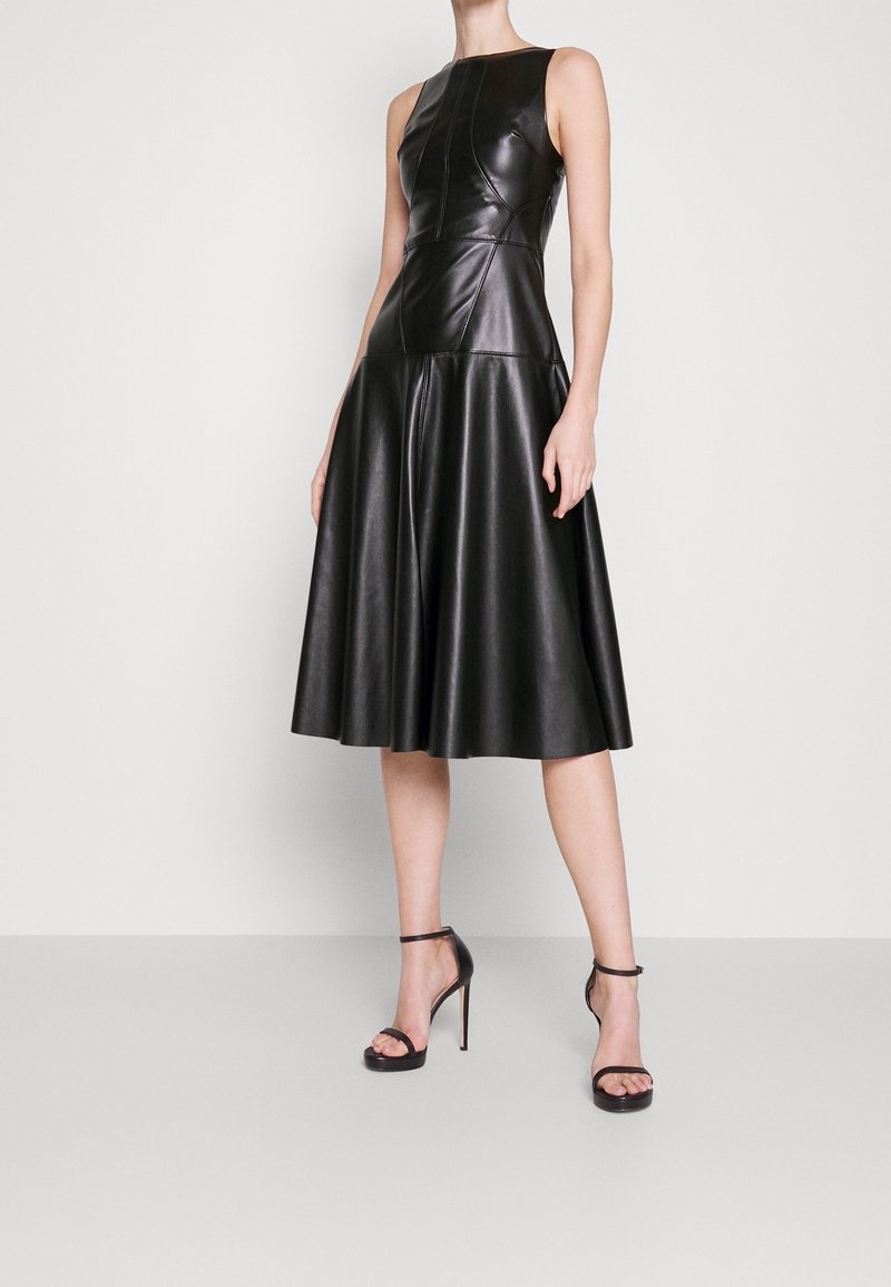 Svart läder ärmlös klänning med figurnära korsett och utställd kjol. Har sömdetaljer och en slät yta. Stylen är kompletterad med svarta högklackade sandaler.