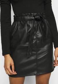 Svart fusk läderkjol med resår i midjan, utrustad med ett knytbälte. Slät textur och subtila sömnadsdetaljer längs fållen.