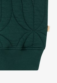 Sötétzöld pulóver anyag, amelyen tűzött mintázat látható kerek formákkal. Bordás mandzsetta az alján. Kis címke a márkanevével látható.