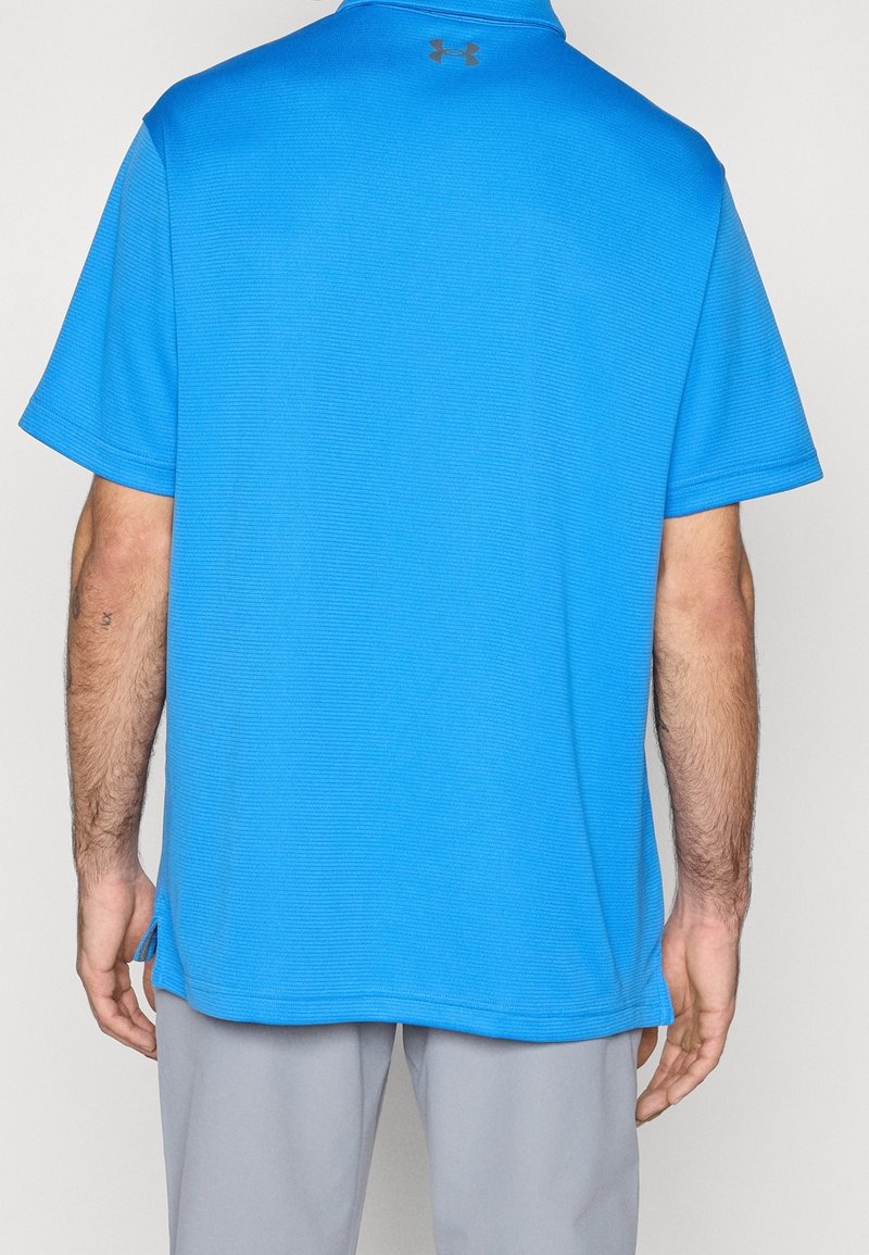 Homme portant un polo bleu vif à manches courtes avec un petit logo noir sur le haut du dos, associé à un pantalon gris clair.