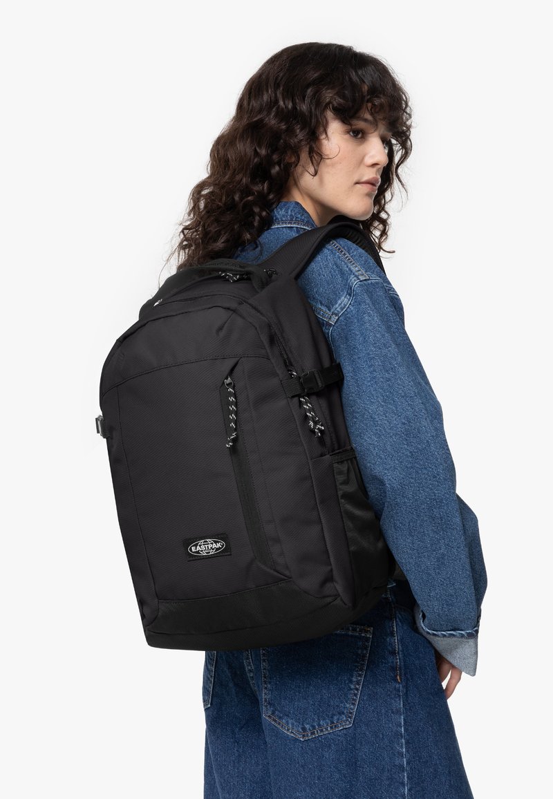 Černý látkový batoh s více přihrádkami, nastavitelnými popruhy a strukturovaným povrchem; má logo a detaily se zipy.