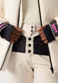 Giacca imbottita bianca con dettagli in nero, chiusura a zip e grande logo rosa. La fascia in vita nera presenta tre bottoni e una texture arricciata.