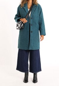 Cappotto turchese con trama, maniche lunghe, bottoni neri, abbinato a pantaloni di jeans scuri a gamba larga e stivaletti neri; borsetta piccola con motivo bianco e nero.
