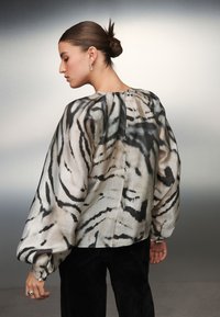 Bluza dugih rukava od mješavine svile, s uzorkom tigrastih pruga u krem i crnoj boji. Nabranih rukava i visokog ovratnika sa zatvaranjem straga.