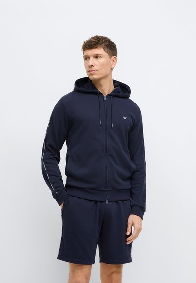 Μπλε σκούρα φούτερ με φερμουάρ και κουκούλα με κορδόνι, με μικρό λογότυπο στο στήθος και κείμενα στο μήκος των μανικιών. Συνδυάζεται με σορτς σε ίδιο χρώμα.
