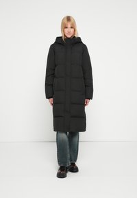 Must pikk sulejope kapuutsiga, tikandiga, esikinnituse ja küljetaskutega, kantud helesiniste teksapükste ja mustade saapade kohal.