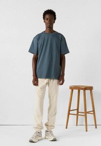 T-shirt blu scuro a maniche corte abbinato a pantaloni jogger beige chiaro e sneakers neutre, su uno sfondo semplice con uno sgabello di legno.