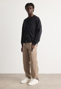 Black cardigan with buttons, textured fabric, and patch logo, paired with light beige pants with zippered pockets and white sneakers.