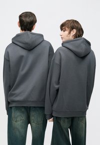 Dvaja jednotlivci nosiaci nadrozmerné tmavosivé mikiny z mäkkej látky, ktoré majú kapucňu, spustené ramená a voľný strih.