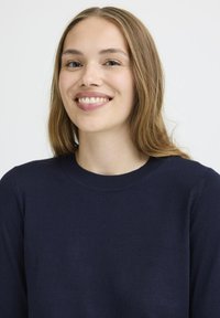Pull en tricot bleu marine avec un col rond, texture lisse et manches ajustées, porté par une personne aux longs cheveux châtain clair.