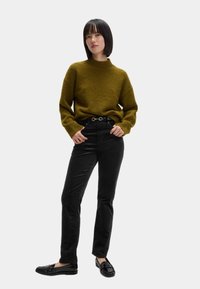 Olivgrüner Strickpullover mit hohem Kragen, kombiniert mit schwarzen geraden Hosen und schwarzen Slippern. Schlichte, aber moderne Silhouette.