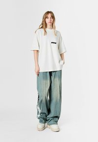 Camiseta blanca de gran tamaño con el logo negro "MWM", combinada con jeans azules desgastados y sueltos que presentan un estampado gráfico y detalles de utilidad, y zapatos beige gruesos.