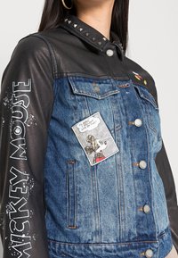 Giacca in denim con maniche in pelle nera, borchie argento sul colletto, stampa bianca di Topolino e una toppa in stile fumetto sul petto.