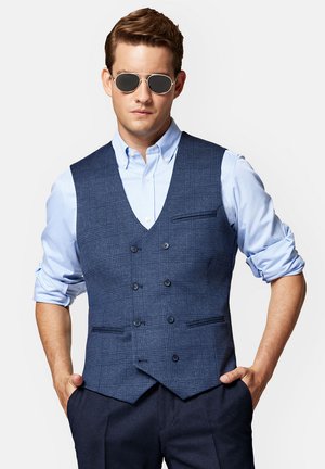Granatowa kamizelka w kratę o slim fit, podwójnie zapinana z przodu, z dwiema kieszeniami. Nosi się ją na jasnoniebieskiej koszuli i w połączeniu z okularami przeciwsłonecznymi.
