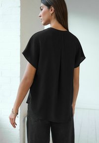 Schwarze kurzärmlige Bluse mit einer Raffung auf der Rückseite. Hergestellt aus einem glatten Stoff, bietet sie eine lockere Passform und einen abgerundeten Saum, kombiniert mit dunklen Jeans.