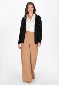 Schwarze Strickjacke mit Paillettenverzierungen an den Ärmeln, kombiniert mit einer weißen Satinbluse und beigen, hoch taillierten, weiten Hosen. Neutrale Absätze.