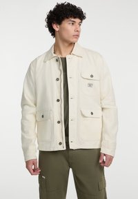 Hellbeige Jacke aus weichem Stoff, mit Kragen, Knopfverschluss, zwei frontalen Taschen und einem kleinen Markenlabel.