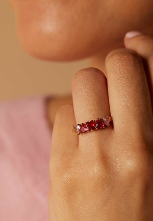 Main portant une bague en or ornée de cinq pierres précieuses en forme de cœur, roses et rouges, tenue près du visage sur un fond beige doux.