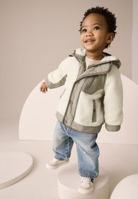 Veste crème et grise doublée en polaire, avec capuche, poches zippées et boutons-pression, associée à un jean bleu clair déchiré et des baskets blanches.