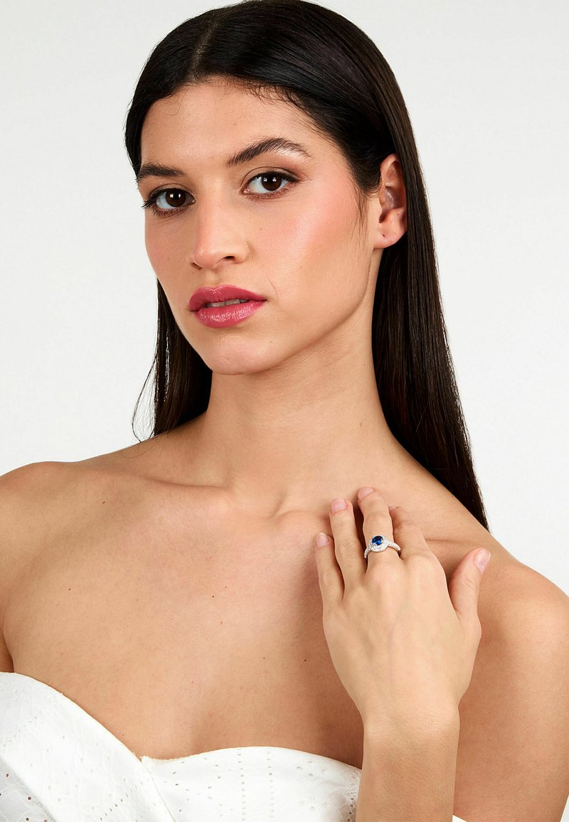 Anello d'argento con pietra blu, caratterizzato da dettagli intricati. Il modello ha i capelli lisci e scuri e indossa un top bianco senza spalline.