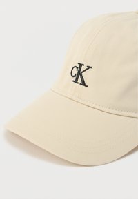 Μπεζ καπέλο του μπέιζμπολ από υφή υφάσματος, με μαύρο κεντημένο λογότυπο "cK" στο μπροστινό μέρος και καμπύλη γείσο.