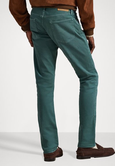 Zaļie pieguļošie džinsi ar gludu faktūru, tajos ir aizmugurējās kabatas un ādas birka uz jostas. Saskaņoti ar brūnām loafers.