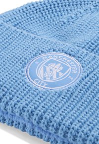 Bonnet tricoté bleu clair avec texture côtelée et une écusson circulaire du logo de Manchester City en bleu clair et blanc à l'avant.