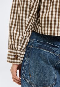 Κοντινό πλάνο ατόμου που φοράει καρό πουκάμισο καφέ και λευκό με κουμπωτό μανίκι και μπλε τζιν τζην με ορατή πίσω τσέπη και ραφές.