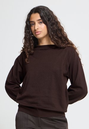Jeune femme aux longs cheveux bouclés portant un pull marron foncé à manches longues et un pantalon gris sur un fond uni.