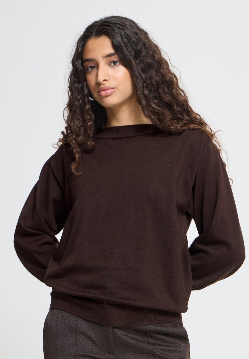 Jeune femme aux longs cheveux bouclés portant un pull marron foncé à manches longues et un pantalon gris sur un fond uni.