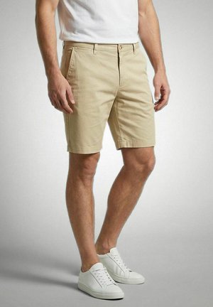 Man draagt beige korte broek, wit T-shirt en witte sneakers, staat voor een effen achtergrond.
