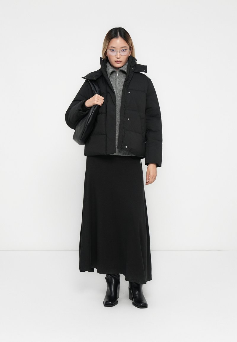 Schwarze gepolsterte Jacke mit hohem Kragen, kombiniert mit einem grauen Rollkragenpullover und einem langen schwarzen Rock. Schwarze Stiefeletten und eine Handtasche.