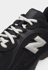 Približni posnetek črnega športnega čevlja z belimi vezalkami in belim logosom "N" z ročnim šivom na strani, ki ima mrežaste in semiš plošče.