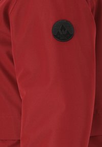 Punainen takki, jossa on sileä pinta ja kuminen logo-ompelus hihassa, jossa on vuoristokuvio ja teksti "Whistler Tanska".