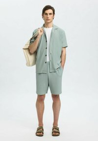 Maglietta a maniche corte verde chiaro, texturizzata, e pantaloni corti abbinati con bottoni e tasche, abbinati a una maglietta bianca e sandali marroni.