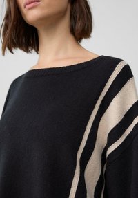 Schwarzer Strickpullover mit rundem Ausschnitt, auf einer Seite mit cremefarbenen und beigen diagonalen Streifen. Weiche Textur, lockere Passform.